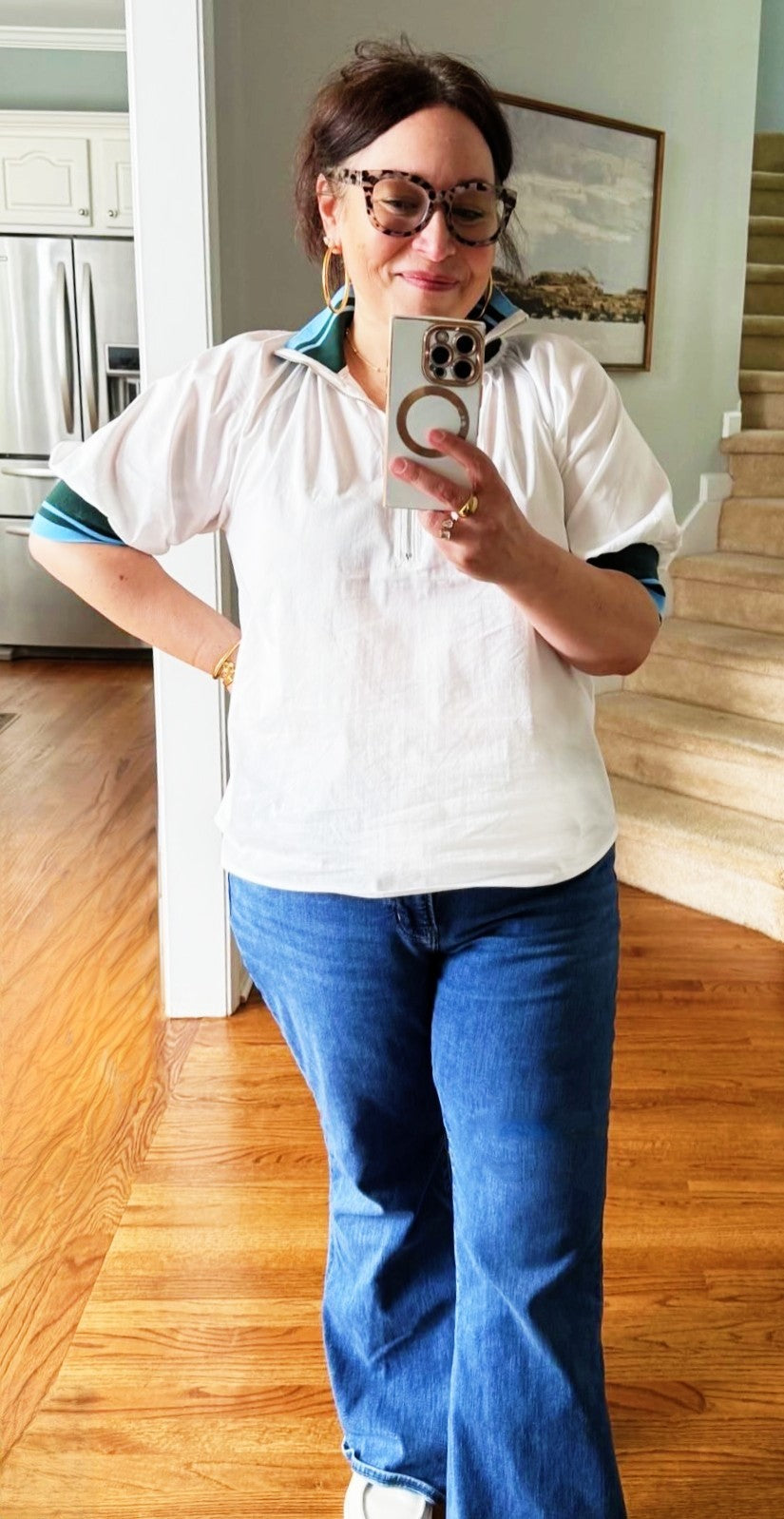 Woman taking a mirror selfie wearing a white shirt with blue accents and blue jeans in a home setting.